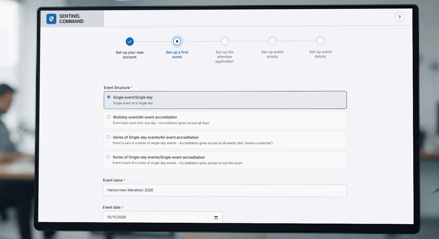 A monitor on an office desk showing event set-up: a five-step progress bar with the account step complete, above a form where the event structure, name and date have been filled in.